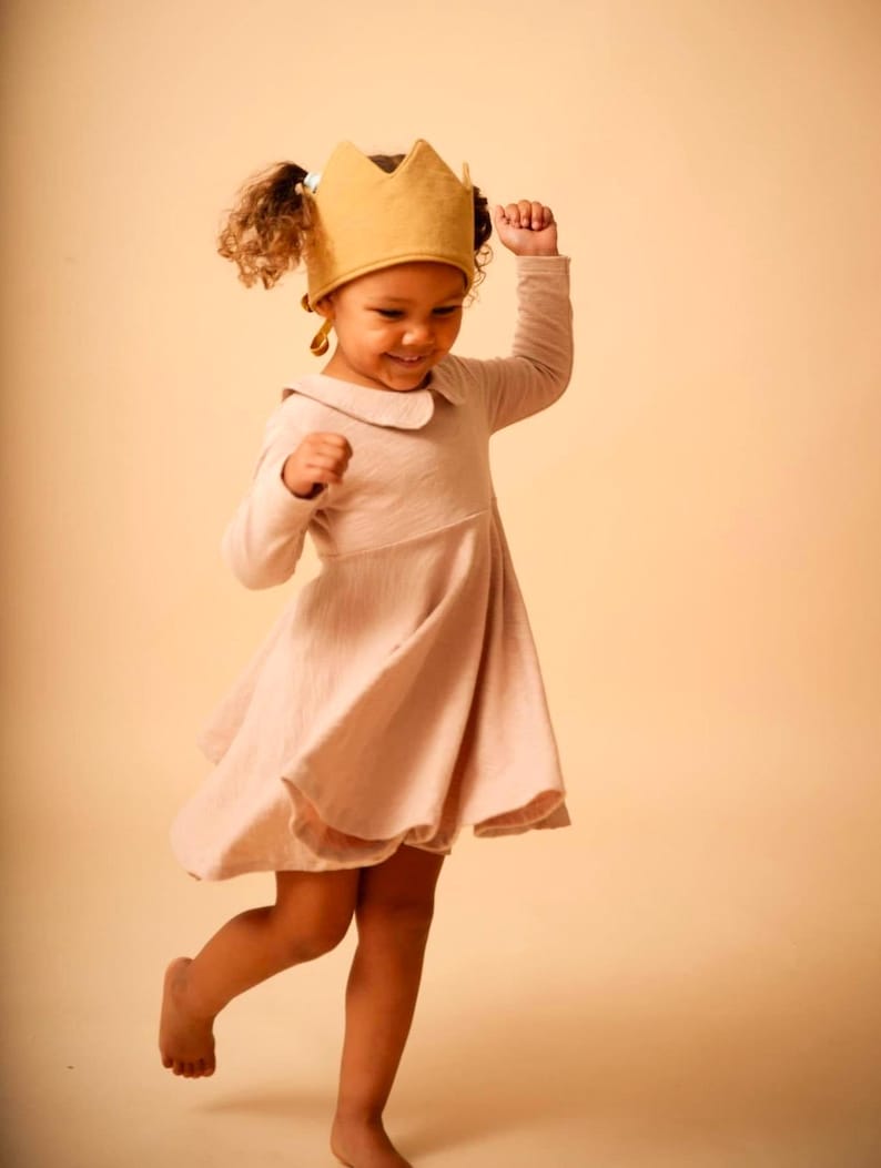 May include: A young girl wearing a pink dress and a gold crown is smiling and twirling.