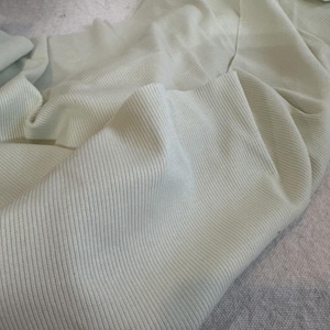 May include: A pale mint green fabric featuring a fine vertical ribbed texture. The material is draped in soft, flowing folds, highlighting the consistent linear pattern and the light, muted color of the textile surface throughout the piece.