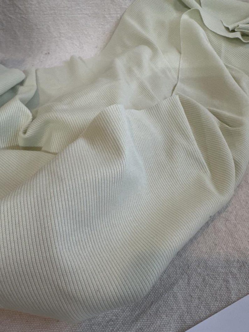 May include: A pale mint green fabric featuring a fine vertical ribbed texture. The material is draped in soft, flowing folds, highlighting the consistent linear pattern and the light, muted color of the textile surface throughout the piece.