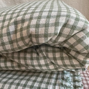 May include: A folded textile featuring a sage green and off-white gingham check pattern. The fabric has a visible woven texture and a frayed, decorative edge detail along the bottom section, showcasing a soft and layered appearance.