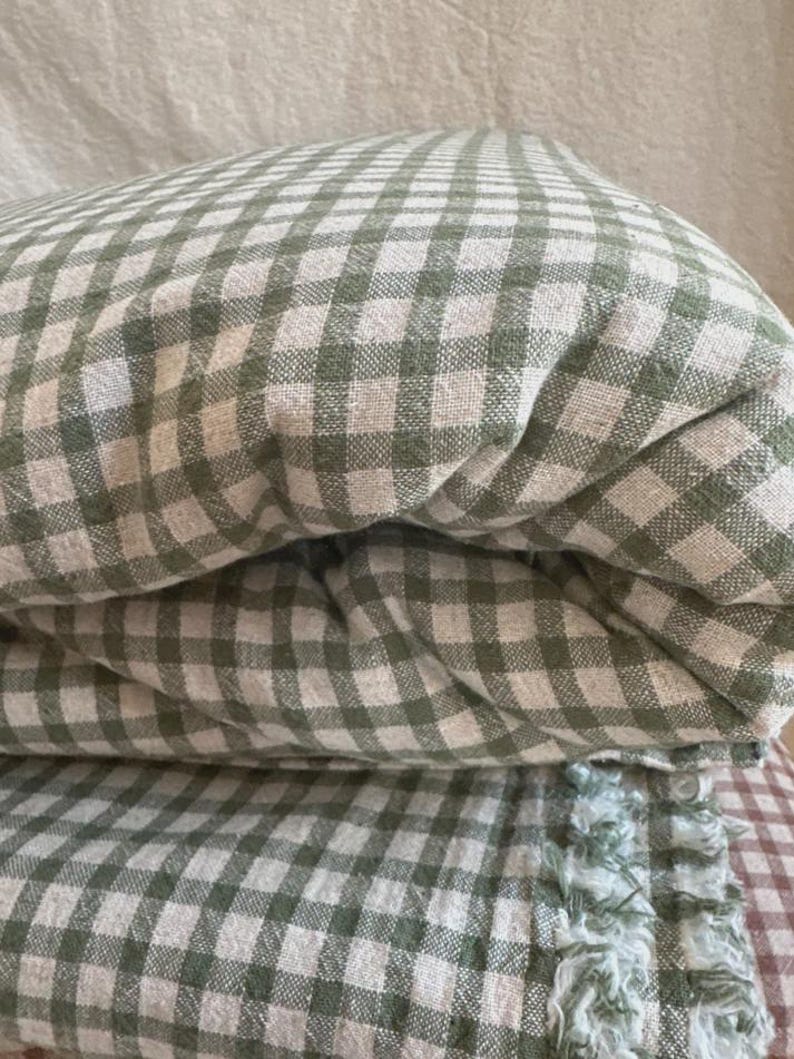 May include: A folded textile featuring a sage green and off-white gingham check pattern. The fabric has a visible woven texture and a frayed, decorative edge detail along the bottom section, showcasing a soft and layered appearance.