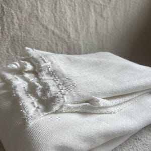 May include: Folded white fabric with a textured surface. The top layer has a decorative, fringed edge. The fabric appears to be soft and possibly linen or cotton. The image is well-lit, highlighting the texture and details.