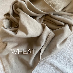 May include: A close-up of a folded, beige fabric with the word "WHEAT" in white block letters. The fabric has a textured appearance and a raw edge. The fabric is draped over a white surface.