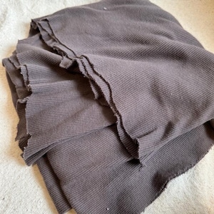 May include: A dark brown fabric with a ribbed texture and raw, unfinished edges. The material is folded in layers, showcasing the vertical ribbed pattern and the frayed, uneven texture along the borders of the dark brown textile piece.