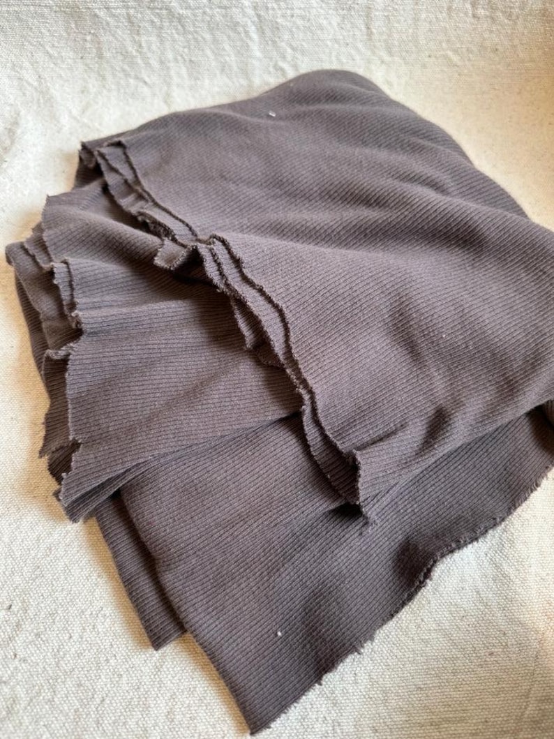May include: A dark brown fabric with a ribbed texture and raw, unfinished edges. The material is folded in layers, showcasing the vertical ribbed pattern and the frayed, uneven texture along the borders of the dark brown textile piece.