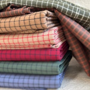 Flat lay of multiple yards of brushed cotton grid plaid fabric in assorted colorways. Each piece shows the fine plaid detailing, with a brushed finish that adds softness and warmth. Great fabric for quilting, crafting, and comfortable clothing. The image highlights the fabric’s texture and versatility for DIY projects or custom-made gifts.