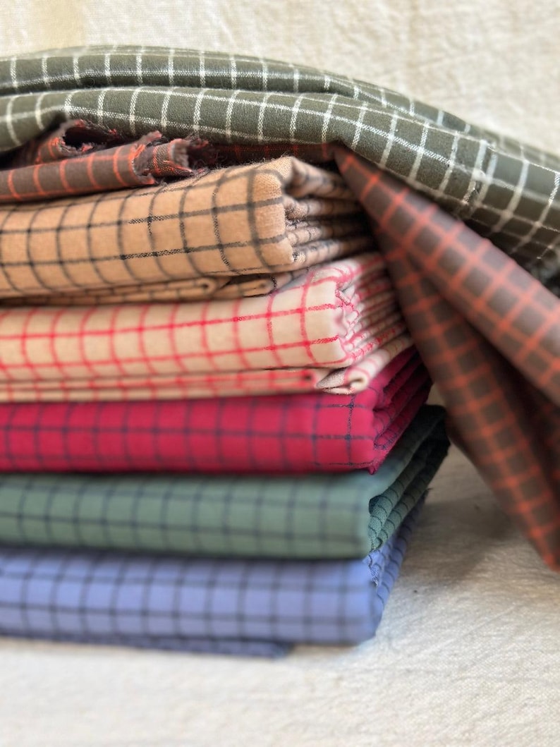 Flat lay of multiple yards of brushed cotton grid plaid fabric in assorted colorways. Each piece shows the fine plaid detailing, with a brushed finish that adds softness and warmth. Great fabric for quilting, crafting, and comfortable clothing. The image highlights the fabric’s texture and versatility for DIY projects or custom-made gifts.