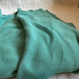 May include: A soft, teal-colored knitted blanket featuring a prominent vertical ribbed texture throughout the fabric. The textile displays a consistent, solid green-blue hue with visible knit rows that create a structured, linear pattern across the surface.