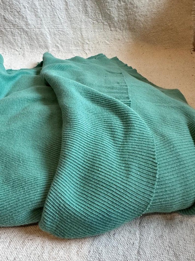 May include: A soft, teal-colored knitted blanket featuring a prominent vertical ribbed texture throughout the fabric. The textile displays a consistent, solid green-blue hue with visible knit rows that create a structured, linear pattern across the surface.