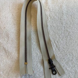 May include: A beige zipper with dark brown teeth and a black pull tab. The zipper is open and curved, showing the teeth and the fabric tape. The zipper is likely for sewing projects or clothing.