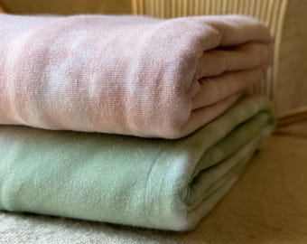 May include: Two folded blankets, one in a soft pink hue and the other in a muted green, are stacked on top of each other. The blankets appear to be made of a soft material, and are neatly folded. The background is a light beige.
