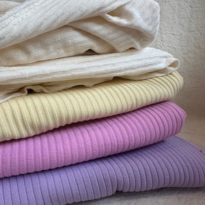 May include: A stack of five folded fabric pieces in various colors. The top two are off-white with a subtle textured grid pattern. The bottom three are solid ribbed knit fabrics in pale yellow, vibrant pink, and soft lavender, respectively.