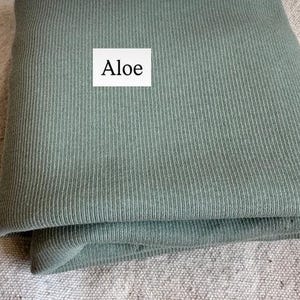 May include: A folded piece of sage green fabric with a vertical ribbed texture. A white rectangular label is placed on the fabric with the word Aloe printed in black text. The material appears soft and has a consistent matte finish throughout.
