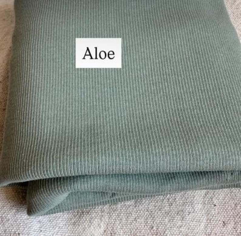 May include: A folded piece of sage green fabric with a vertical ribbed texture. A white rectangular label is placed on the fabric with the word Aloe printed in black text. The material appears soft and has a consistent matte finish throughout.
