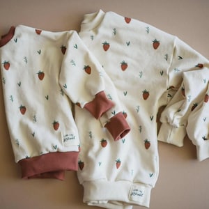 May include: Two long-sleeve sweatshirts in an off-white color featuring a repeating pattern of small red strawberries and green sprigs. Both garments have solid dark red ribbed cuffs and necklines, with a small white label reading Long Live Girlhood.