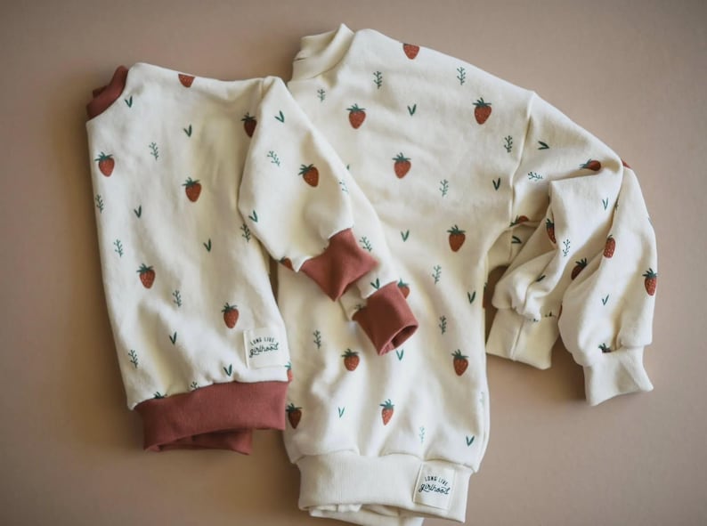 May include: Two long-sleeve sweatshirts in an off-white color featuring a repeating pattern of small red strawberries and green sprigs. Both garments have solid dark red ribbed cuffs and necklines, with a small white label reading Long Live Girlhood.