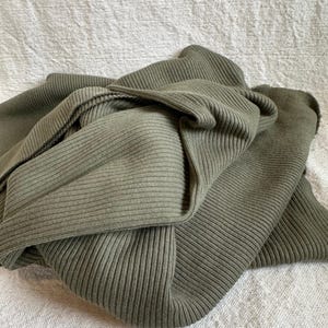 May include: A folded piece of olive green fabric featuring a distinct vertical ribbed texture. The material is draped in soft, overlapping layers, showcasing the consistent linear pattern and the muted, earthy tone of the textile throughout the folds.