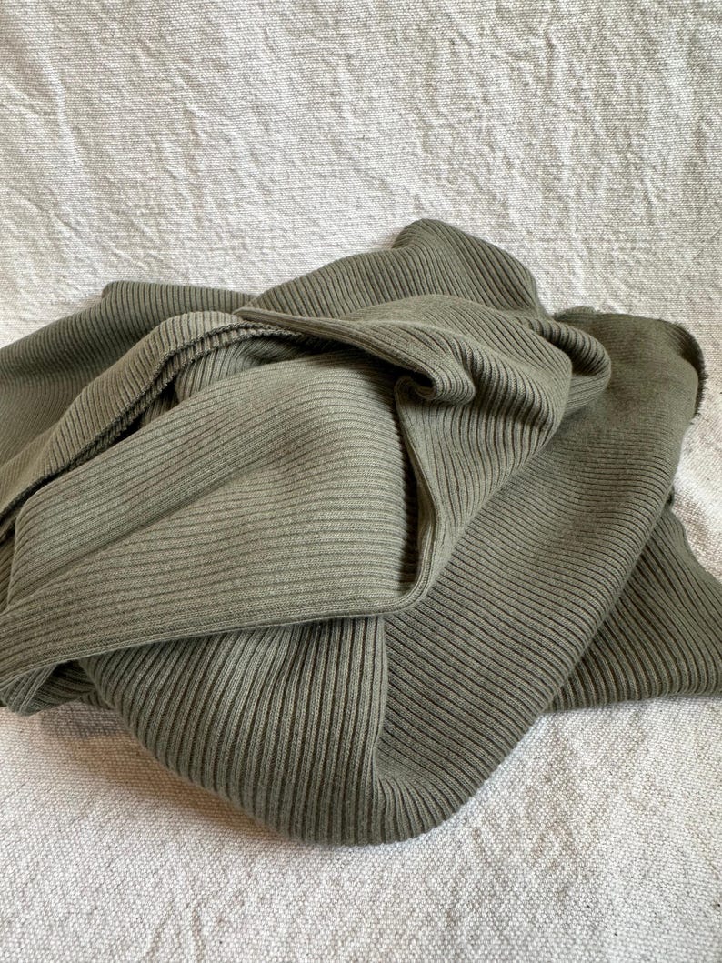 May include: A folded piece of olive green fabric featuring a distinct vertical ribbed texture. The material is draped in soft, overlapping layers, showcasing the consistent linear pattern and the muted, earthy tone of the textile throughout the folds.