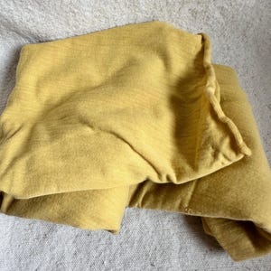 May include: A folded, mustard-yellow textile, possibly a blanket or sheet, rests on a textured, light grey surface. The fabric appears soft and has a subtle, horizontal weave pattern. The edges of the textile are slightly ruffled.