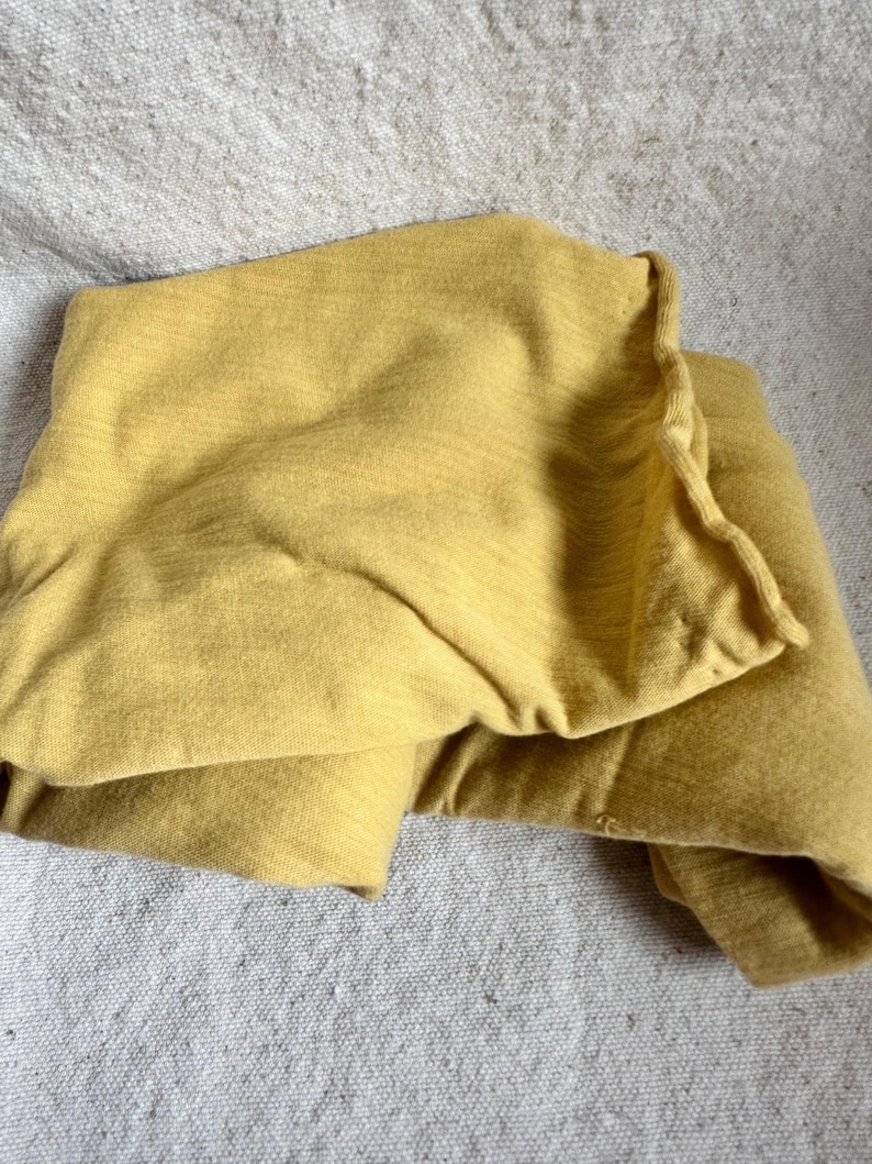May include: A folded, mustard-yellow textile, possibly a blanket or sheet, rests on a textured, light grey surface. The fabric appears soft and has a subtle, horizontal weave pattern. The edges of the textile are slightly ruffled.