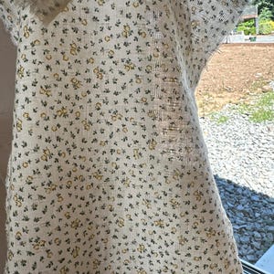 May include: A piece of off-white fabric featuring a repeating pattern of tiny yellow flowers and small green leaves. The textile has a visible woven grid texture and frayed edges, showing a semi-sheer quality when held up to the light.