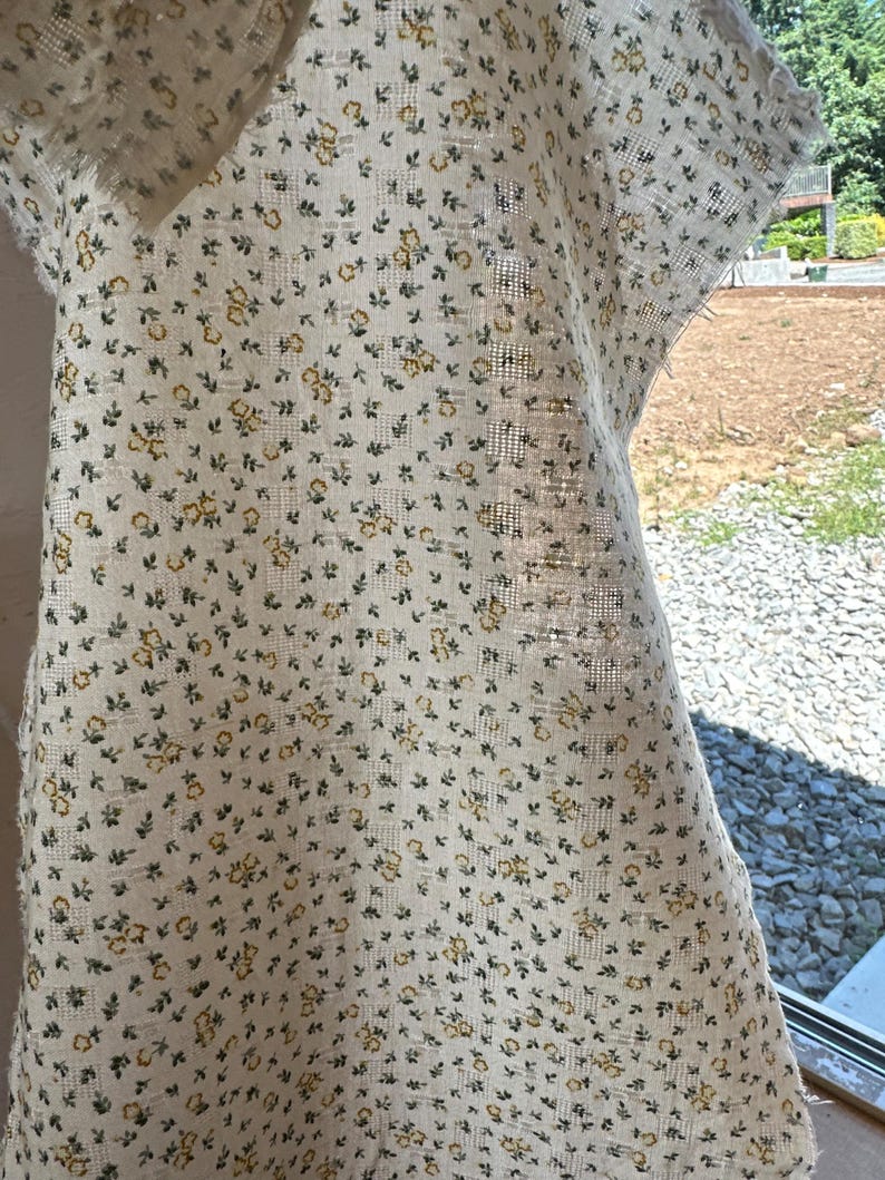 May include: A piece of off-white fabric featuring a repeating pattern of tiny yellow flowers and small green leaves. The textile has a visible woven grid texture and frayed edges, showing a semi-sheer quality when held up to the light.