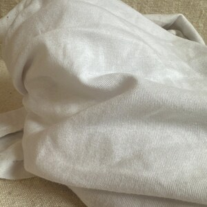 May include: A plain white t-shirt made of a soft, lightweight knit fabric. The garment is shown in a folded, bunched-up state, highlighting the fine texture of the material and the consistent, solid white color throughout the entire piece of clothing.
