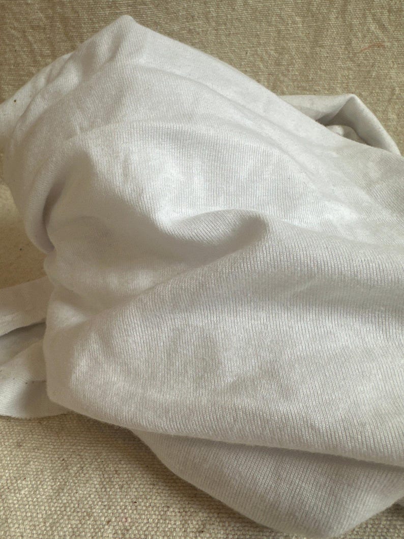 May include: A plain white t-shirt made of a soft, lightweight knit fabric. The garment is shown in a folded, bunched-up state, highlighting the fine texture of the material and the consistent, solid white color throughout the entire piece of clothing.