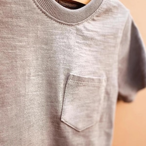 May include: A grey t-shirt with a chest pocket. The short-sleeved top has a crew neck and is hanging on a wooden hanger. The fabric has a textured appearance.