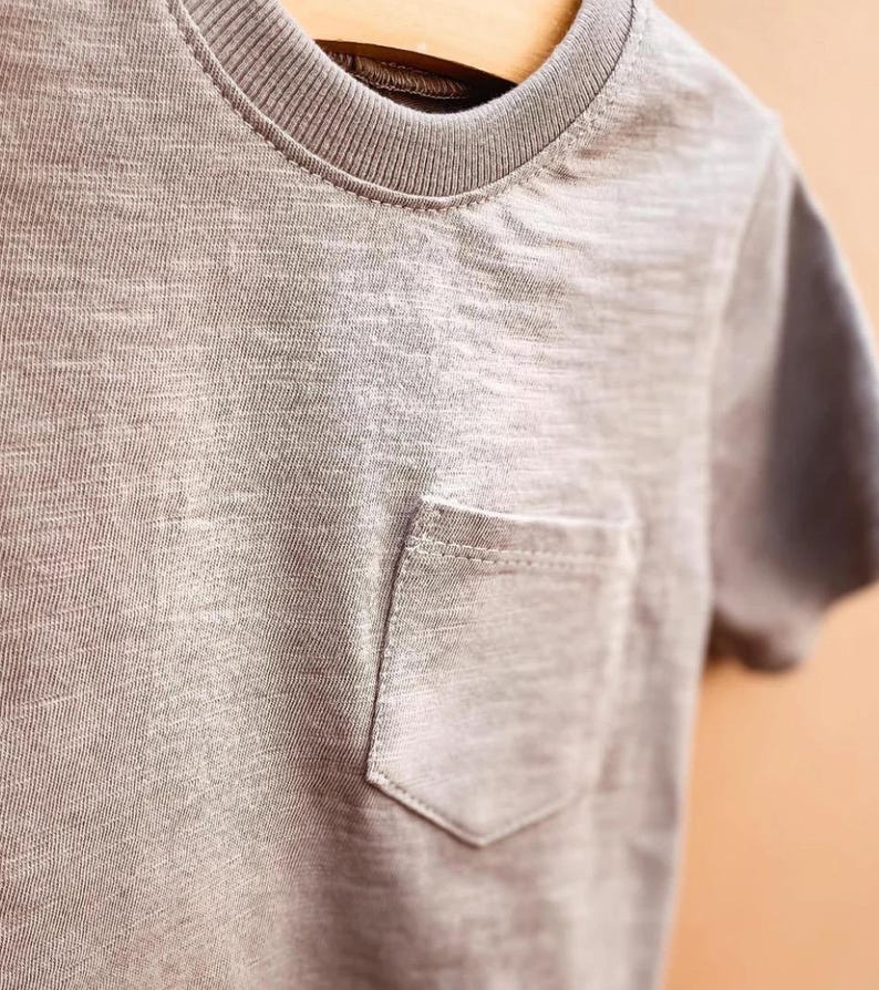 May include: A grey t-shirt with a chest pocket. The short-sleeved top has a crew neck and is hanging on a wooden hanger. The fabric has a textured appearance.