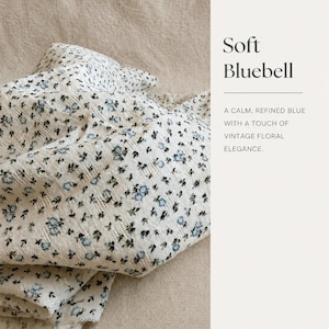May include: A white fabric featuring a small-scale blue and black floral pattern with a textured weave. The text on the right side reads: Soft Bluebell, A CALM, REFINED BLUE WITH A TOUCH OF VINTAGE FLORAL ELEGANCE.