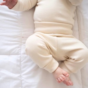 May include: A cream-colored long-sleeve shirt and matching pants set made of a ribbed knit fabric. The top features long sleeves and a fitted waistband, while the pants have cuffed ankles, providing a simple and uniform appearance in light beige.