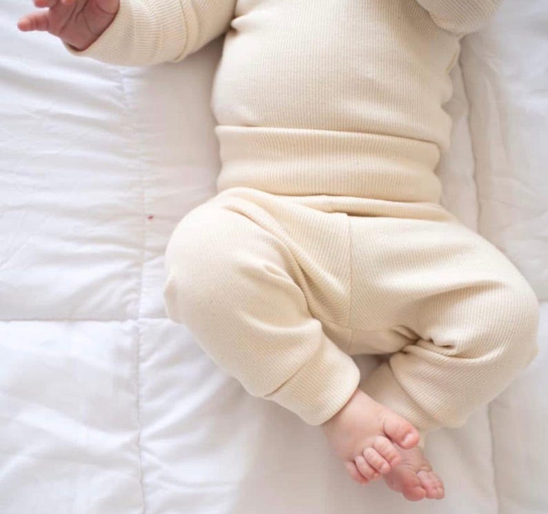 May include: A cream-colored long-sleeve shirt and matching pants set made of a ribbed knit fabric. The top features long sleeves and a fitted waistband, while the pants have cuffed ankles, providing a simple and uniform appearance in light beige.