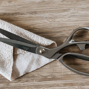 May include: A pair of silver fabric shears with a black handle resting on a beige fabric. The shears have the text "INFERNO" and "J.L. 1980" printed on the blade.
