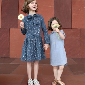 Puede incluir: Dos niñas con vestidos tomadas de la mano, de pie frente a una pared de ladrillos rojos. Una niña lleva un vestido azul con motas blancas y zapatos de lunares blancos y negros. La otra niña lleva un vestido azul claro y sandalias doradas.