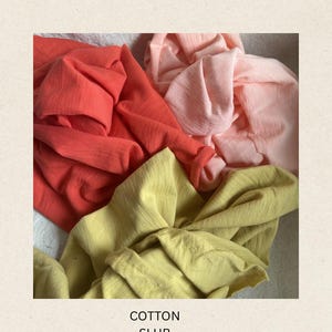 May include: Close-up of four swatches of cotton fabric in shades of coral, peach, and yellow-green. The fabric is textured and draped, with the words "COTTON SLUB" printed at the bottom.