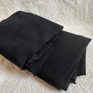 May include: A folded piece of black fabric featuring a distinct vertical ribbed texture. The material has raw, unfinished edges along the folds, showcasing the knit construction of the textile. The fabric is layered, creating depth and shadows.
