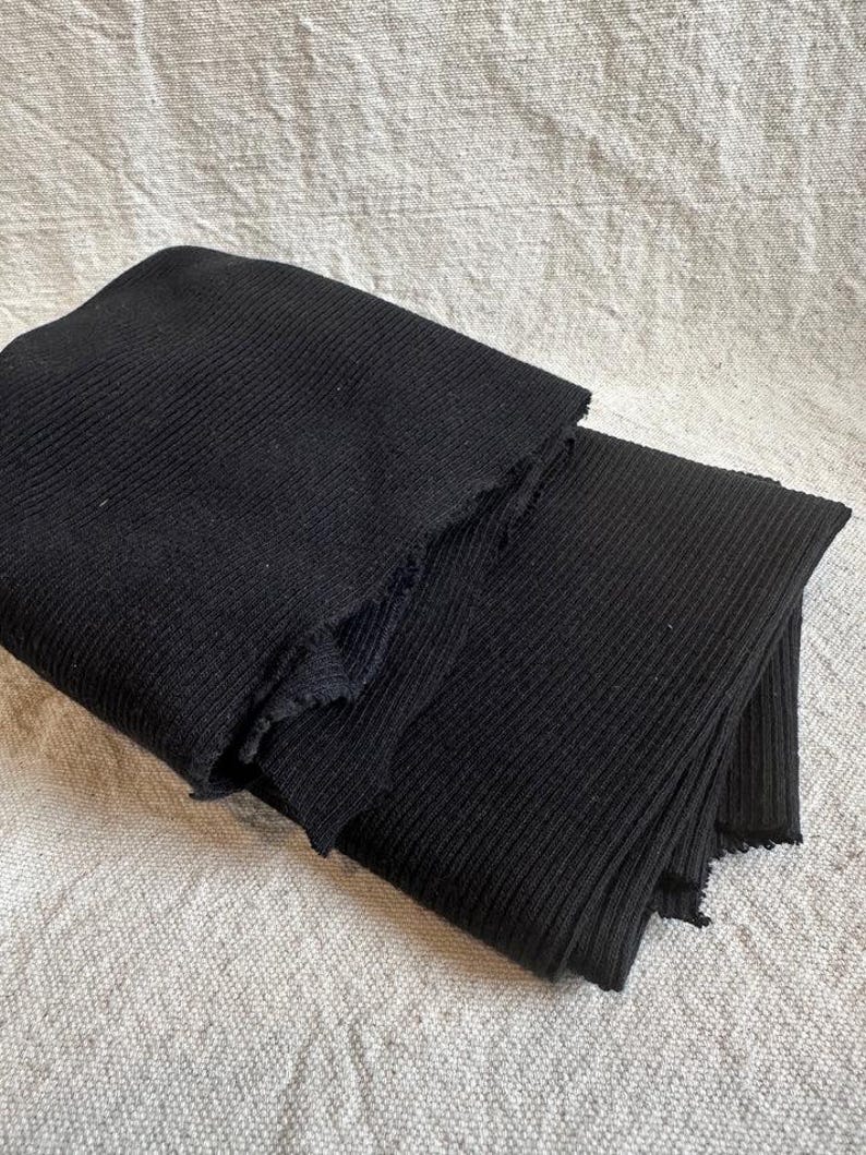 May include: A folded piece of black fabric featuring a distinct vertical ribbed texture. The material has raw, unfinished edges along the folds, showcasing the knit construction of the textile. The fabric is layered, creating depth and shadows.