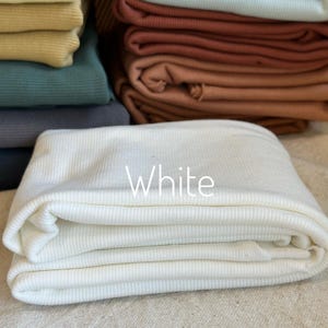 May include: A stack of folded ribbed fabric in various colors including white, light blue, terracotta, dark green, and yellow. The white fabric is in the foreground with the word White printed in white text on the center of the folded material.