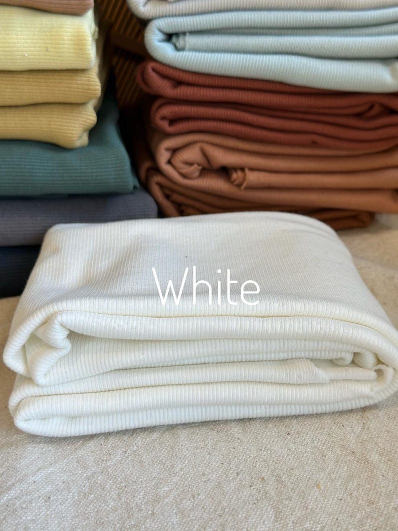 May include: A stack of folded ribbed fabric in various colors including white, light blue, terracotta, dark green, and yellow. The white fabric is in the foreground with the word White printed in white text on the center of the folded material.