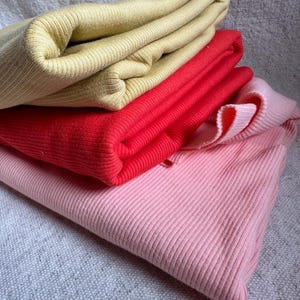 May include: A stack of three folded ribbed knit garments in solid colors. The top piece is a muted mustard yellow, the middle piece is a vibrant cherry red, and the bottom piece is a soft pastel pink. Each item features a consistent vertical ribbed texture.