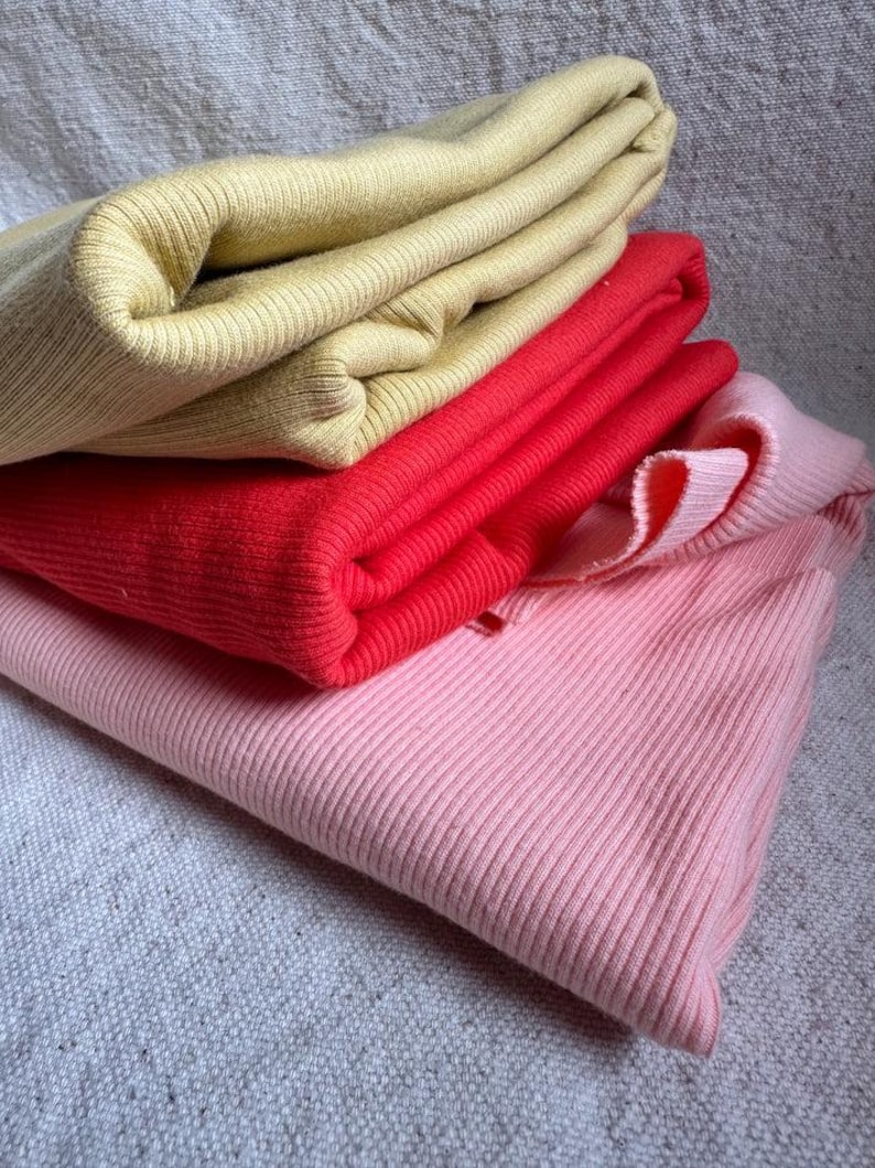 May include: A stack of three folded ribbed knit garments in solid colors. The top piece is a muted mustard yellow, the middle piece is a vibrant cherry red, and the bottom piece is a soft pastel pink. Each item features a consistent vertical ribbed texture.