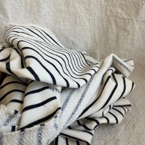 May include: Close-up of a folded fabric with a white base and bold black horizontal stripes. The fabric appears to be a soft knit or terry cloth, with visible texture and a slightly frayed edge. The stripes vary in width.