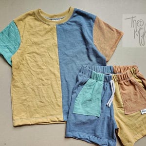 May include: A two-piece children's outfit comprising a short-sleeved t-shirt and matching shorts. The t-shirt is colour-blocked with mustard yellow, teal, and blue panels. The shorts have a blue and mustard yellow colour-block design with a teal pocket.