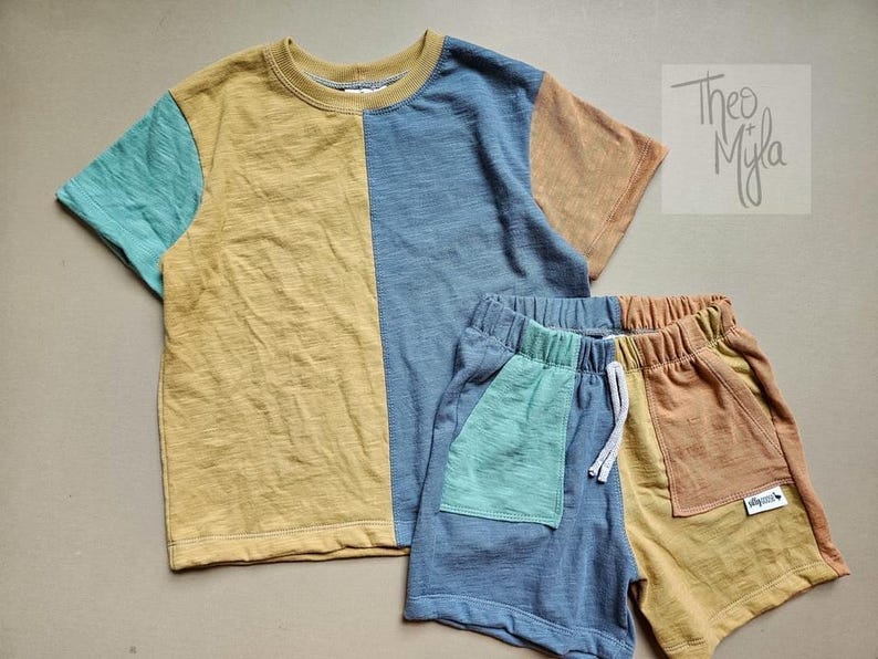May include: A two-piece children's outfit comprising a short-sleeved t-shirt and matching shorts. The t-shirt is colour-blocked with mustard yellow, teal, and blue panels. The shorts have a blue and mustard yellow colour-block design with a teal pocket.