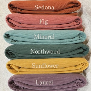 May include: A stack of six folded ribbed fabric pieces in various colors. The colors are labeled with white text as Sedona, Fig, Mineral, Northwood, Sunflower, and Laurel. Each piece of fabric features a consistent vertical ribbed texture.