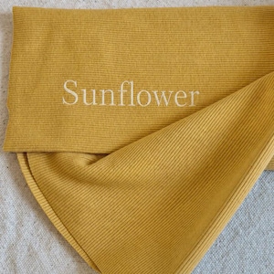 May include: A mustard yellow ribbed fabric accessory featuring the word Sunflower embroidered in white thread. The material has a distinct vertical ribbed texture and is folded over to display the embroidered text clearly on the front surface.