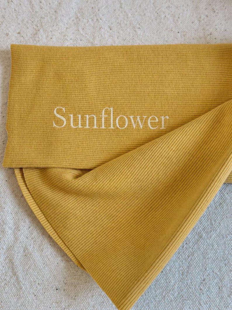 May include: A mustard yellow ribbed fabric accessory featuring the word Sunflower embroidered in white thread. The material has a distinct vertical ribbed texture and is folded over to display the embroidered text clearly on the front surface.