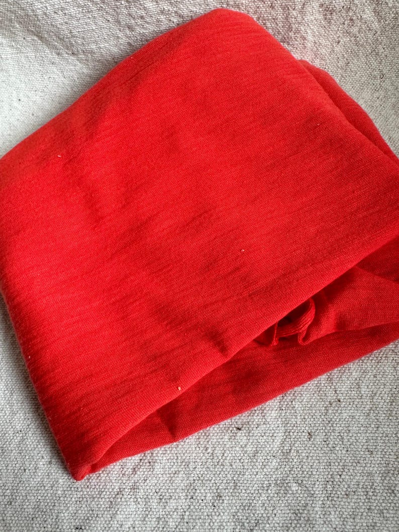 May include: A folded piece of bright red fabric, possibly a scarf or clothing item, with a textured surface. The fabric is neatly folded, highlighting its vibrant colour and potential use in fashion or textiles.