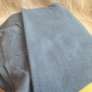 May include: A folded piece of dusty blue fabric with a visible knit texture. The material has a soft, matte appearance with subtle variations in color across the surface, showing a raw, unfinished edge on the left side of the textile.
